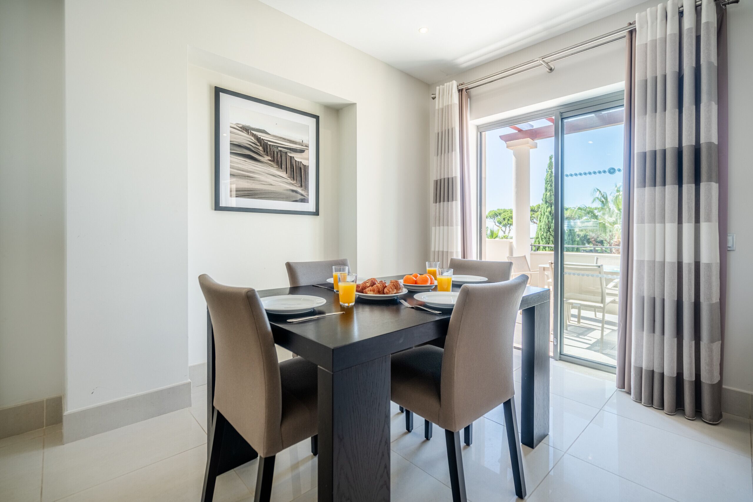 A dining area with a table set for two, a framed picture on the wall, and a view of the outdoors through a glass door.