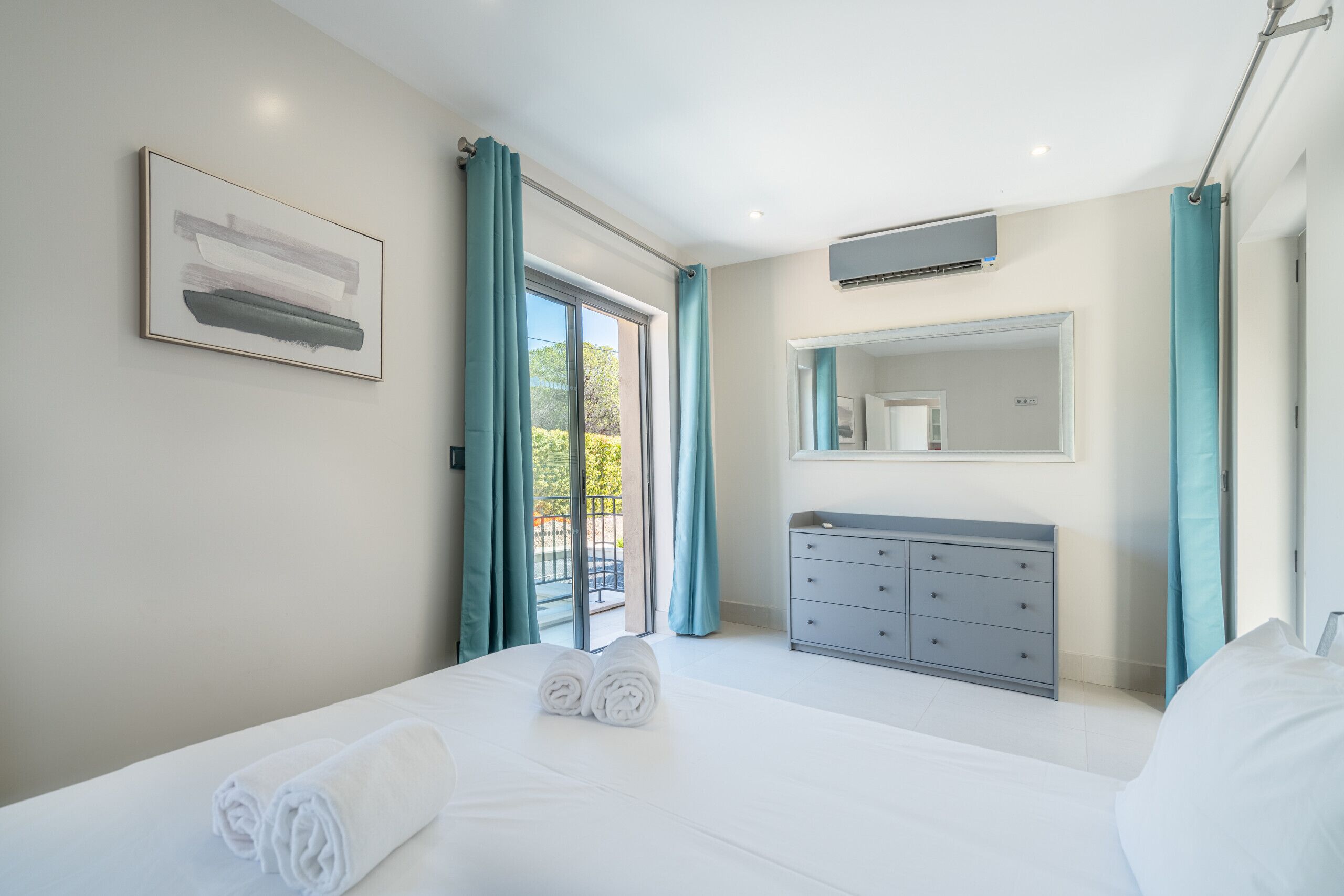 A modern hotel room with a large bed, a grey dresser, a bathroom mirror, and a window with curtains.