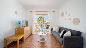 A living room with a grey sofa, a wooden side table, a TV, and a view of a pool and palm trees through a glass door. - Albufeira Palm Tree With Pool by Homing (Albufeira)
