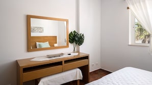 A bedroom with a wooden dresser, a bedside table, a bed, a large mirror, and a window with curtains. - Albufeira Palm Tree With Pool by Homing (Albufeira)