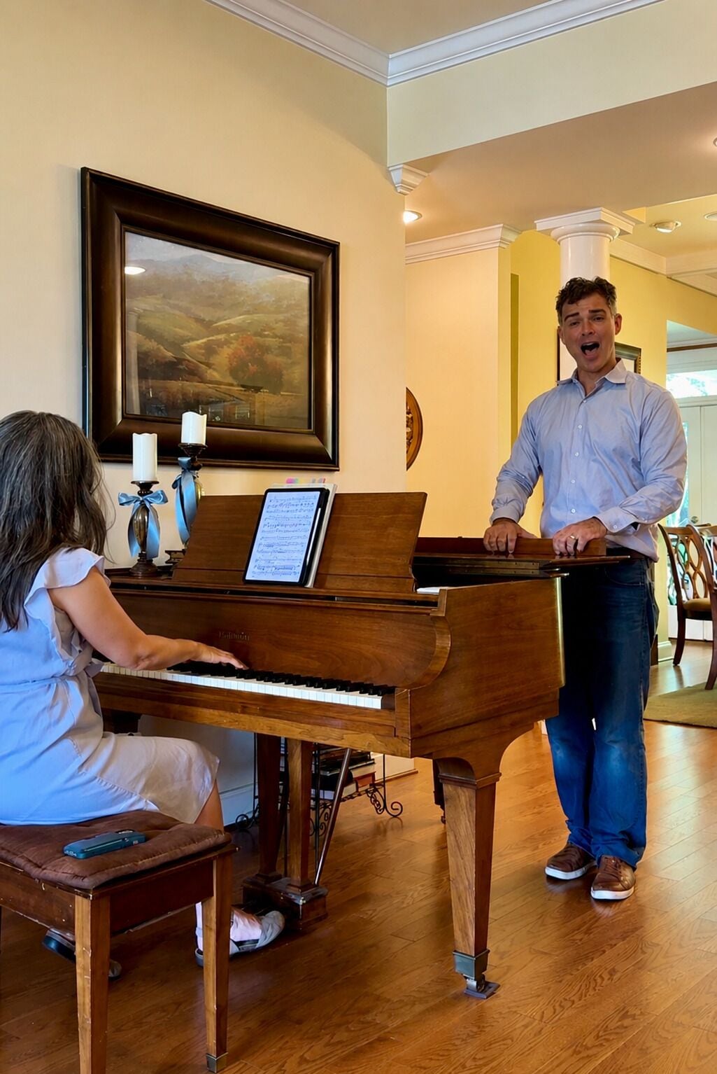 Un piano con partituras, un cuadro de paisaje enmarco y una persona de pie junto a él.