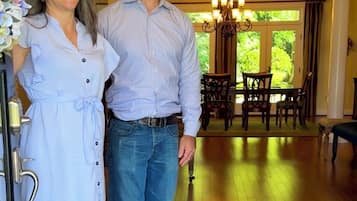 A couple standing in a room with wooden flooring, a chandelier, and a dining area with chairs and a table.