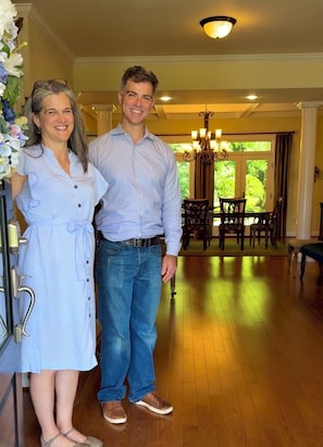 A couple standing in a room with wooden flooring, a chandelier, and a dining area with chairs and a table.