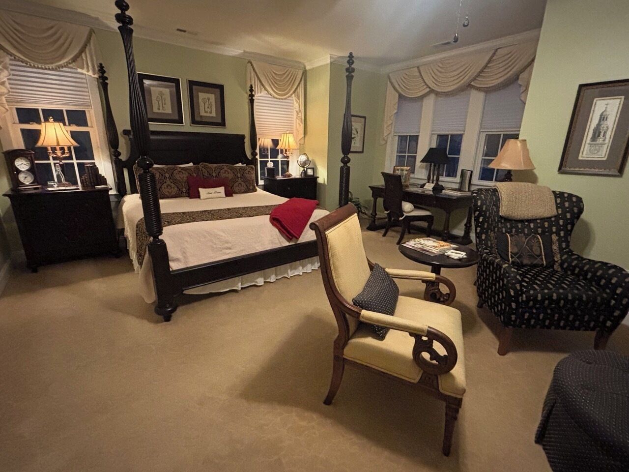 A bedroom with a four-poster bed, a sitting chair, a small table, and a patterned armchair.