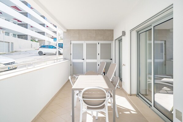 A narrow balcony with a table and chairs, a view of the street, and a building with multiple floors and balconies.