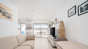 Ein modernes Wohnzimmer mit einer Couch, einem Fernseher und einem kleinen Tisch. Ein Balkon bietet einen Blick ins Freie.