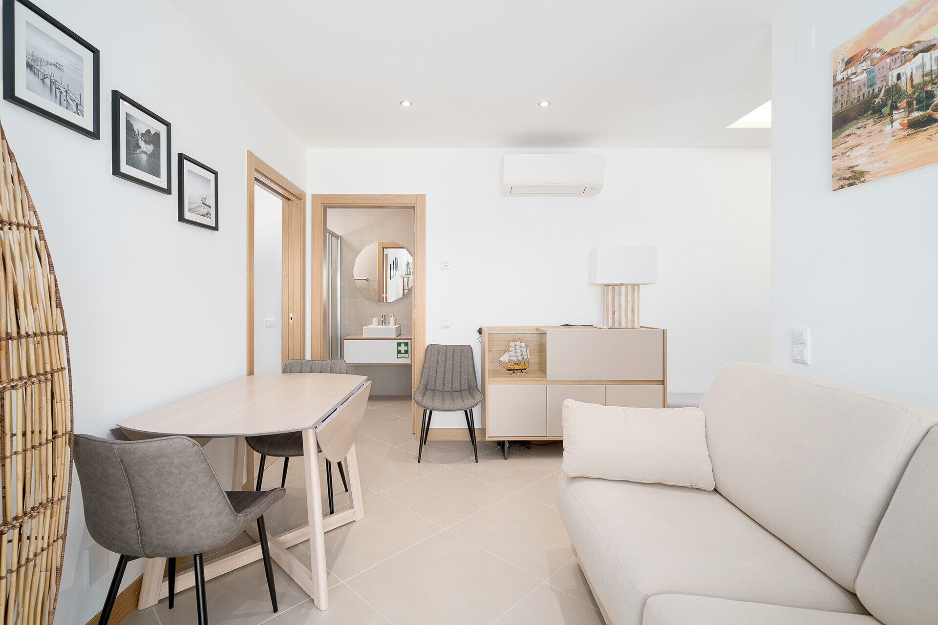 A modern living room with a sofa, a dining table with chairs, a small cabinet, and a wall-mounted air conditioner.