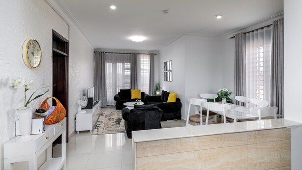 A modern living room with a sofa, coffee table, and dining area. There is a white console table with a vase and a basket. - Elegant Apartment with Stunning Nature views. (Kampala)