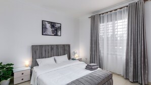 A modern bedroom with a grey upholstered bed, a bedside table with a lamp, a bedside plant, and a large window with sheer curtains. - Elegant Apartment with Stunning Nature views. (Kampala)