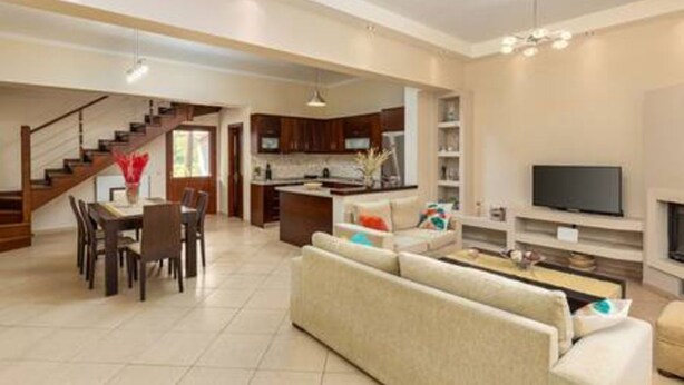 A spacious living area with a sofa, dining table, and chairs, a kitchen with dark cabinets, and a TV mounted on the wall.
