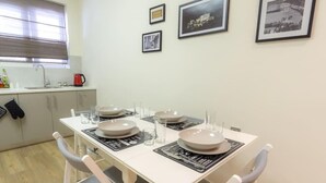 A dining area with a white table set for four, including plates, glasses, and cutlery. There are framed pictures on the wall, a window with blinds, and a kitchen area in the background. - Panathenaic Stadium Apartment by Greece Apartments (Athens)