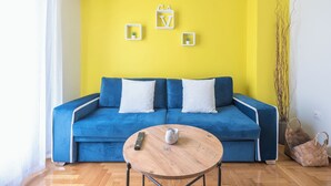Ein hellgelbes Zimmer mit einem blauen Sofa, zwei weißen Kissen, einer Fernbedienung und einer kleinen Pflanze an der Wand.