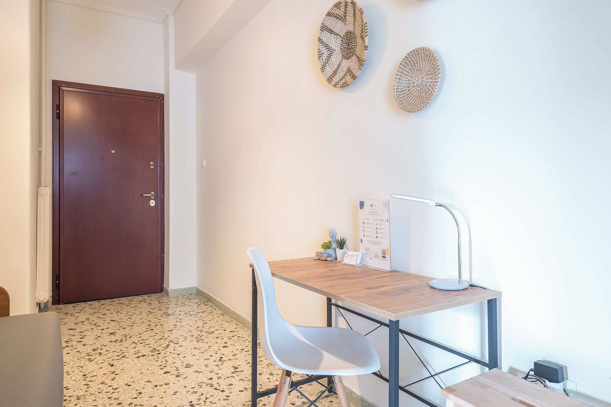 A small, modern room with a wooden desk, a white chair, and a wall-mounted lamp. There is a door to the left and a small table with a plant and a framed certificate on the right.