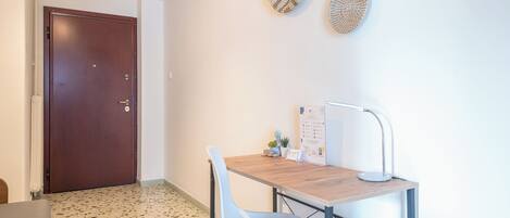 A small, modern room with a wooden desk, a white chair, and a wall-mounted lamp. There is a door to the left and a small table with a plant and a framed certificate on the right.