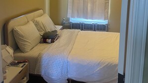 A bedroom with a bed, a bedside table with a lamp, a window with curtains, and a radiator. - Monthly rental - 1-BDR apt, ALL included (hot water, hydro and heating). (Ottawa)