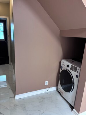 Een wasserette met een wasmachine, een deur naar een andere kamer, en een betegelde vloer.