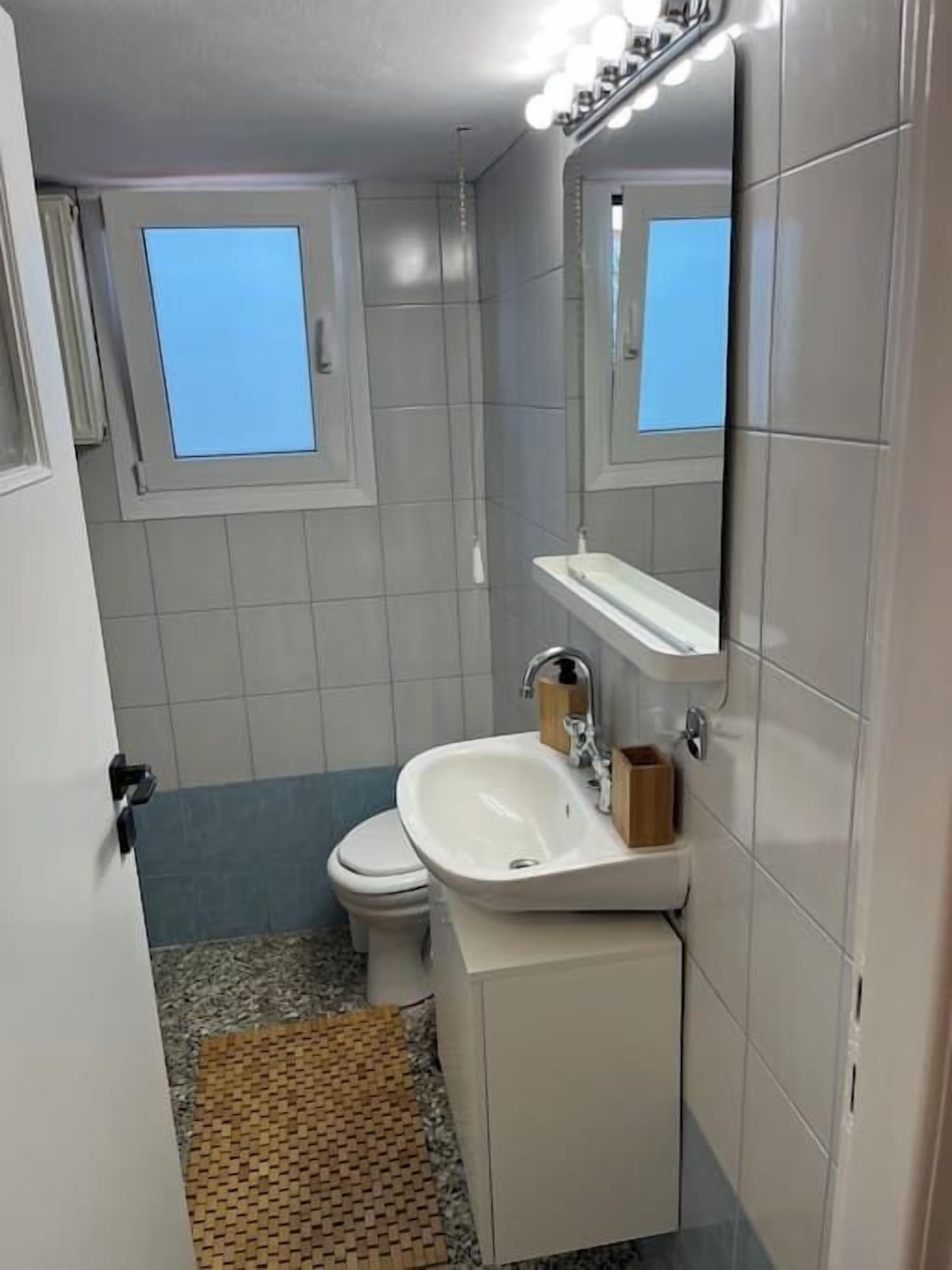 A small bathroom with a toilet, a sink, and a mirror. There is a window above the toilet and a small shelf above the sink.