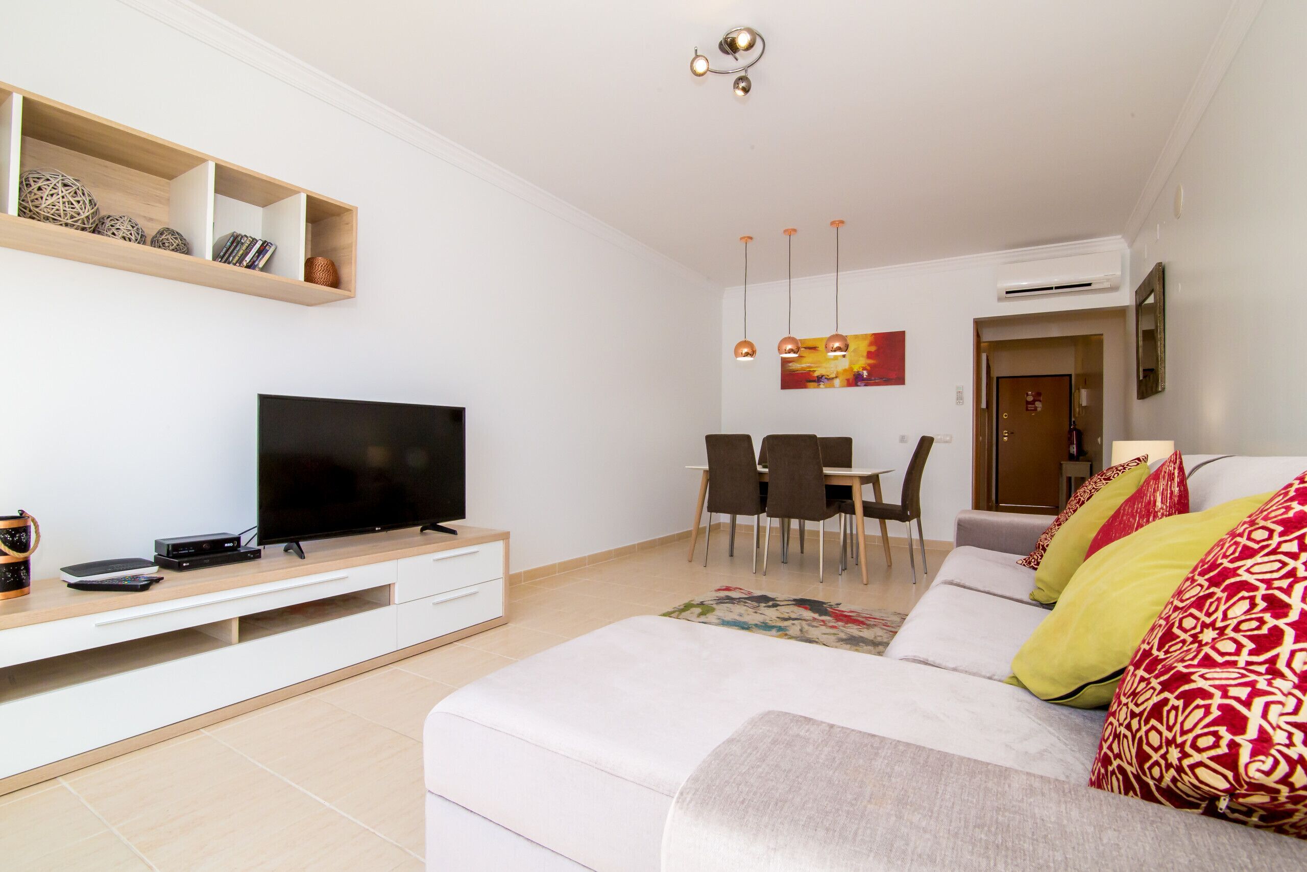 Ein modernes Wohnzimmer mit Flachbildfernseher, Holzregalen, einer Essecke mit Stühlen und einem Sofa mit gemusterten Kissen.