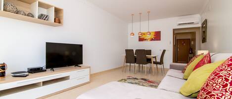 A modern living room with a flat-screen TV, wooden shelves, a dining area with chairs, and a sofa with patterned cushions.