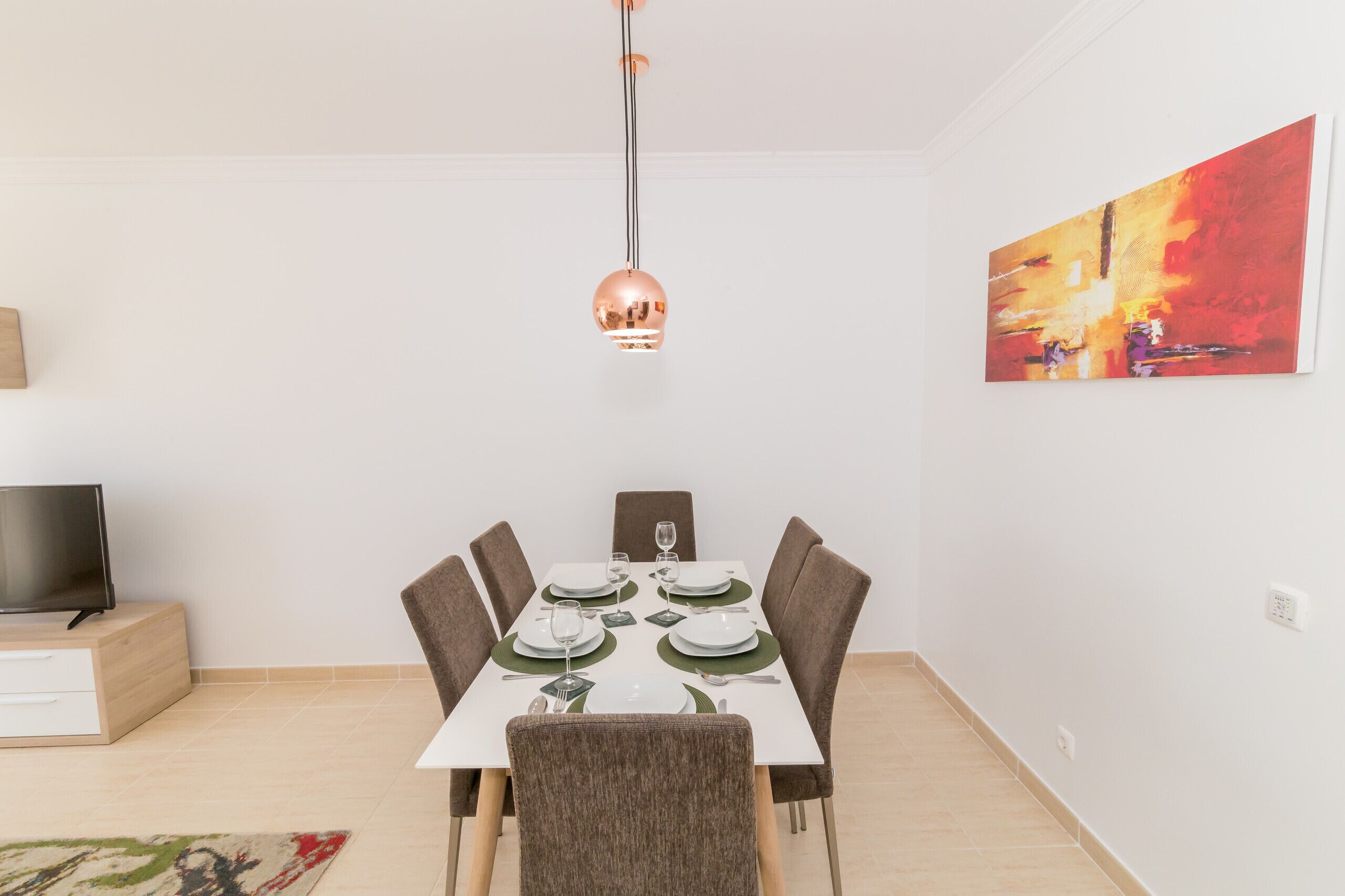 A dining area with a table set for four, a television on a stand, and a painting on the wall.