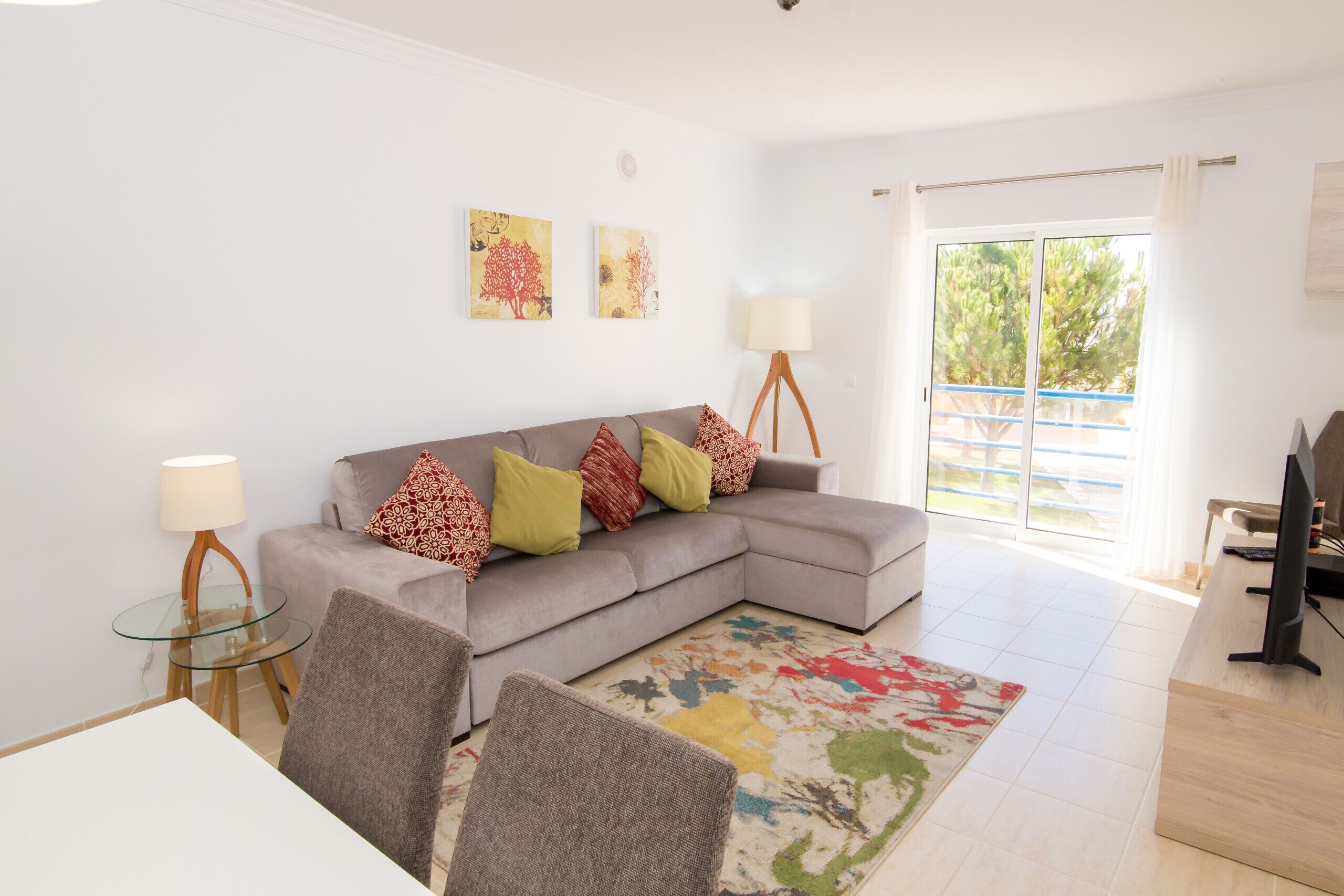 A modern living room with a grey sectional sofa, a glass coffee table, and a colorful rug. There is a TV on a wooden stand, and the room has a large window with a view of trees.