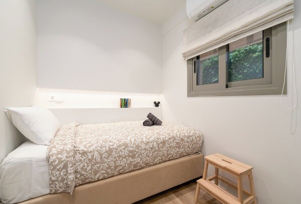 A bedroom with a bed, a bedside table, a window with a view of greenery, and a shelf with books. - Alkis Luxury By Greece Apartments (Zografou)