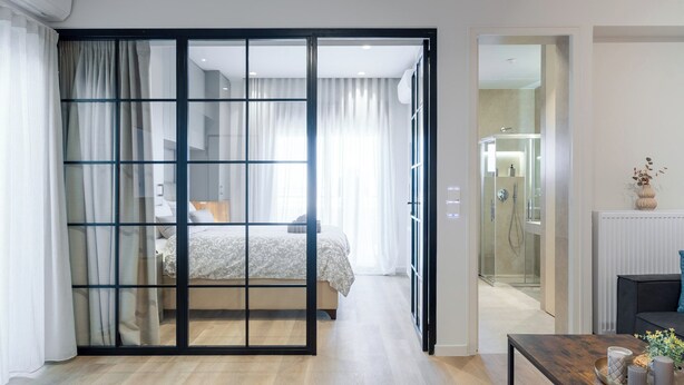 A modern hotel room with a wooden floor, a glass partition separating the living area from the bedroom, a dining table with a vase and books, and a bathroom with a glass shower. - Alkis Luxury By Greece Apartments (Zografou)