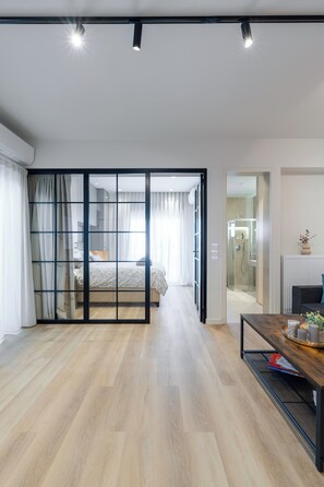 A modern hotel room with a wooden floor, a glass partition separating the living area from the bedroom, a dining table with a vase and books, and a bathroom with a glass shower. - Alkis Luxury By Greece Apartments (Zografou)