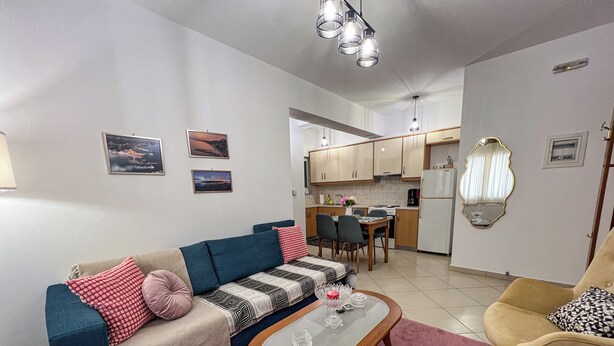 A living room with a sofa, coffee table, and armchair, a kitchen in the background, and a dining area.