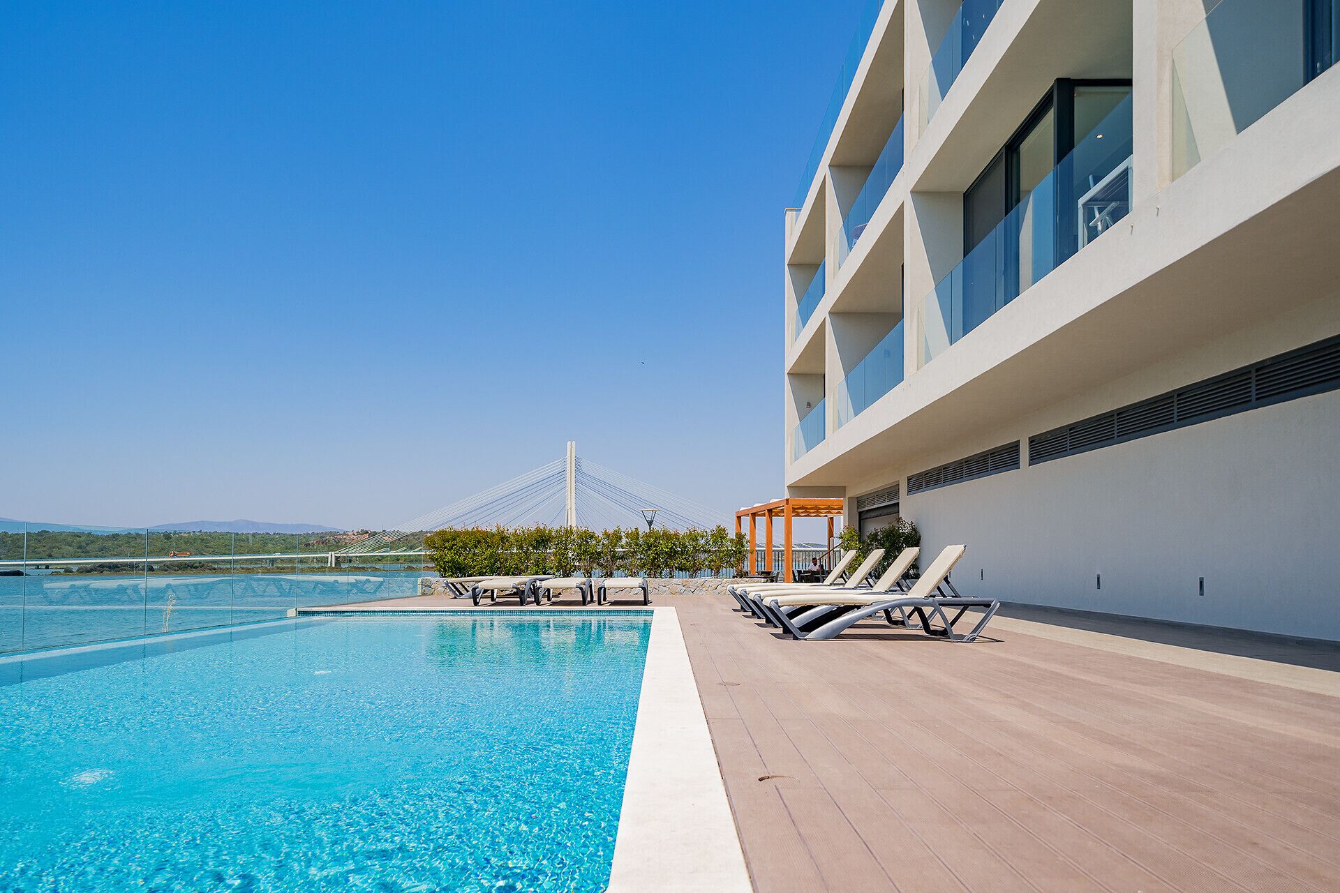 A modern hotel with a rooftop pool, lounge chairs, and a view of a bridge in the background.