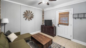 A living room with a ceiling fan, a TV on a wooden cabinet, a green sofa, and a patterned rug. - Serene Midtown Cottage - Walk to Hotspots! (Mobile)