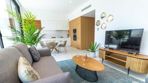 A modern living room with a sofa, coffee table, and TV. There is a kitchen in the background with cabinets and a dining area. - Lagos Deluxe 2 With Pool by Homing (Lagos)