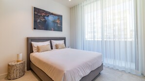 A modern bedroom with a large bed, a nightstand, a painting, and sheer curtains. - Lagos Deluxe 2 With Pool by Homing (Lagos)