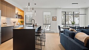 A modern kitchen with a central island, bar stools, and a dining area with a sofa and table. - Ostrya 203 Ski-in/Ski-Out, Steps from the Slopes! (Mont-Tremblant)
