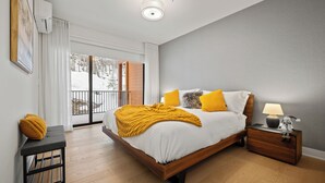 A modern bedroom with a large bed, a nightstand, a bench, and a view of a snowy landscape through the window. - Ostrya 203 Ski-in/Ski-Out, Steps from the Slopes! (Mont-Tremblant)