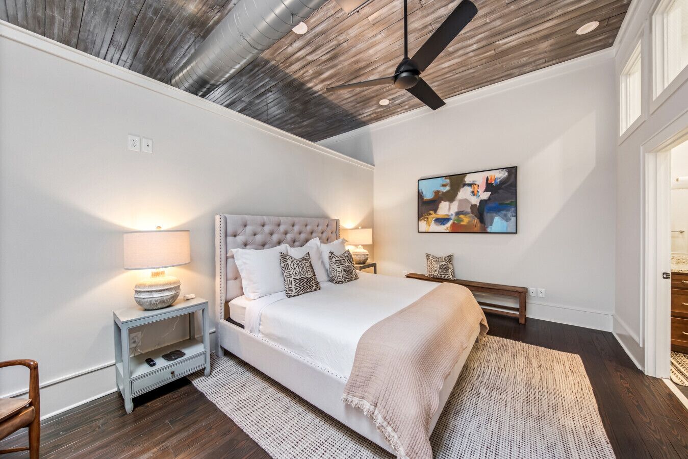 A bedroom with a tufted headboard, a bed with patterned pillows, a bedside table with a lamp, a bench, and a painting on the wall.
