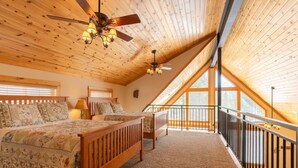 Un dormitorio con techo inclinado, vigas de madera, una cama con colcha estampada, una mesita de noche, una lámpara y vistas a través de una ventana.