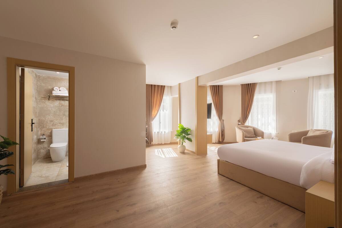 Una habitación de hotel moderna con suelo de madera, una cama grande, una zona de estar con una silla y un baño visible a través de una puerta abierta.