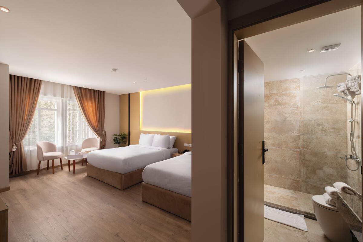 Una habitación de hotel moderna con dos camas, una zona de estar con sillas, una mesita y un área de ducha.