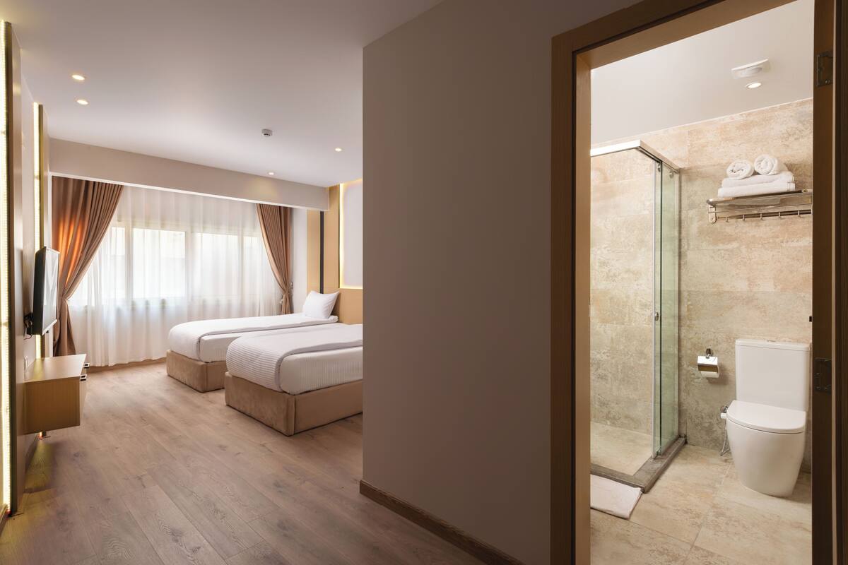 Habitación de hotel con piso de madera, dos camas, un televisor y un baño con ducha de vidrio e inodoro.