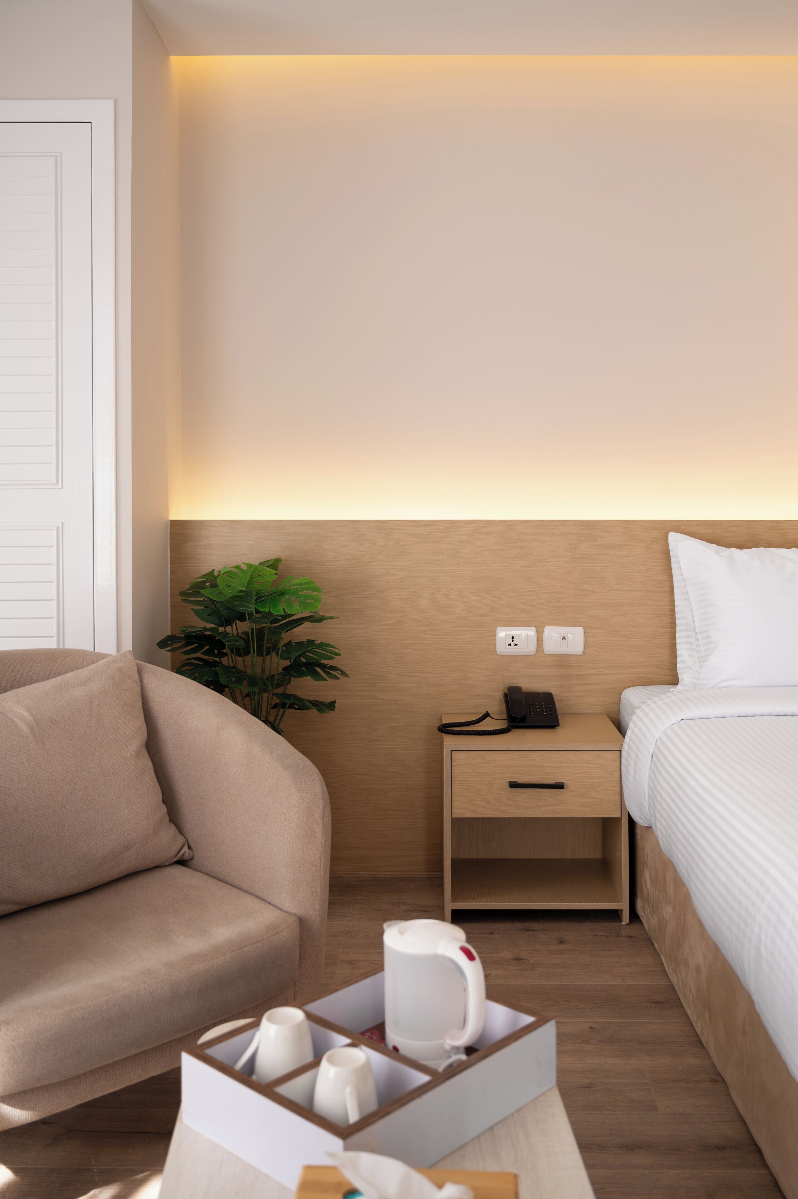 A modern hotel room with a beige sofa, a bedside table with a phone, a bedside lamp, and a small plant.