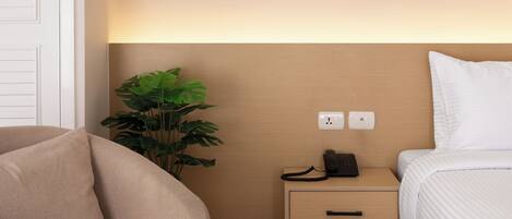 A modern hotel room with a beige sofa, a bedside table with a phone, a bedside lamp, and a small plant.