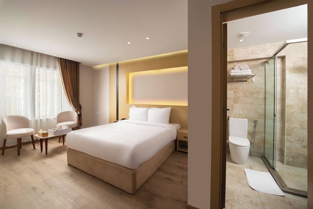Una habitación de hotel con una cama grande, dos sillas, una mesita de noche, cabina de ducha de vidrio y baño con inodoro y lavabo.