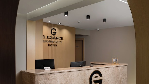 Een hotel receptiebalie met een marmeren aanrecht, een bordje 'Niet roken' en een bord dat 'Elegance Grand City Hotel' vermeldt.