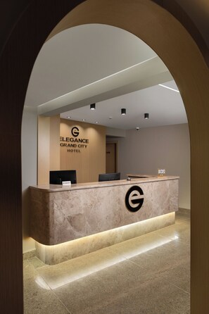 Ein Hotelrezeptionstresen mit Marmorplatte, ein Rauchverbot-Schild und ein Schild mit der Aufschrift 'Elegance Grand City Hotel'.