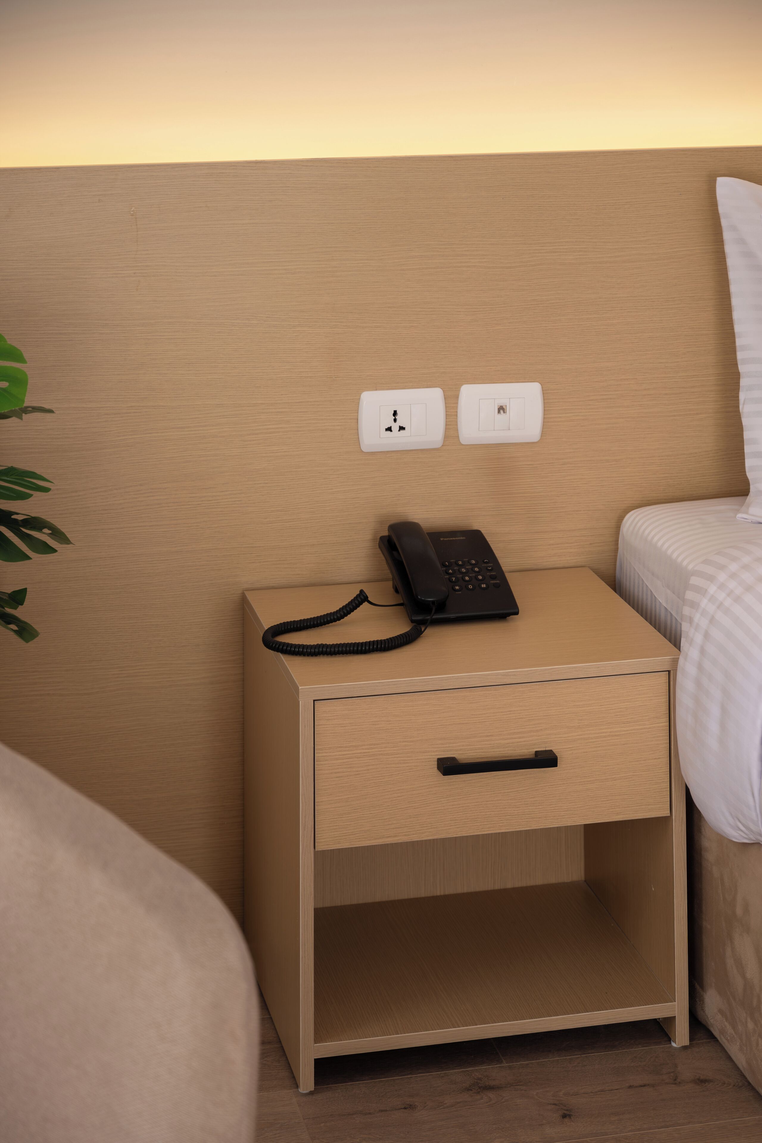 Une table de chevet en bois, avec un téléphone noir, un tiroir et deux prises électriques sur le mur.