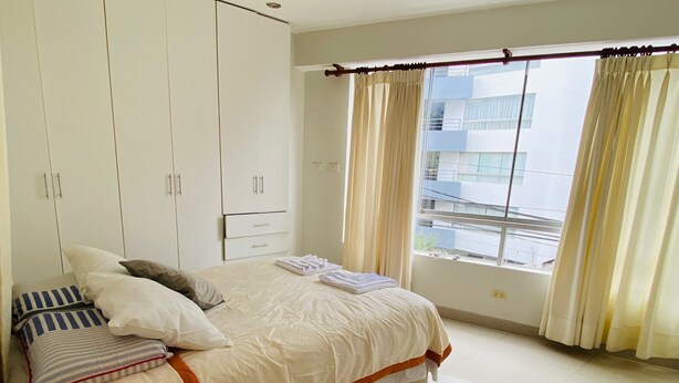 A bedroom with a bed, a nightstand, a lamp, a wardrobe, and a window with curtains. - Refined 3BR Duplex with Ocean Charm (Lima)