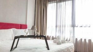 A neatly made bed with a white comforter, a red headboard, a black bedside table with a lamp, and a window with sheer curtains. - I Soho at I City by My Trip & My Stay (Shah Alam)
