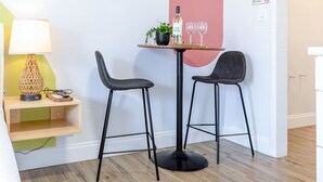 Un petit coin repas avec deux tabourets de bar noirs, une table ronde, une bouteille de vin, deux verres, une plante en pot et une lampe posée sur une étagère en bois.