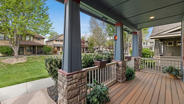 Une véranda couverte avec des piliers en pierre, une terrasse en bois et une vue sur les maisons voisines.